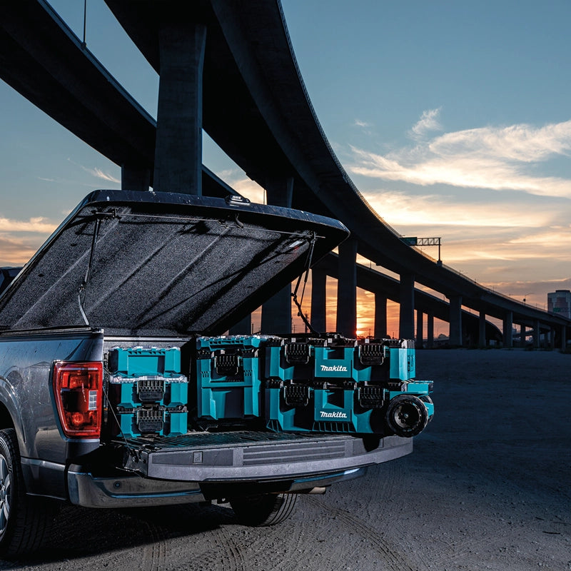 Makita MAKTRAK™ modular storage boxes loaded in a truck bed at a jobsite during sunset under freeway overpasses.

