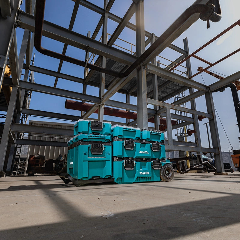 Makita MAKTRAK modular storage system stacked on a dolly at a steel construction jobsite under a clear blue sky.

