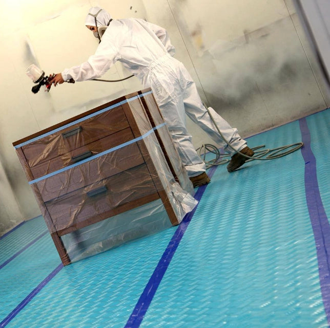 Worker spraying furniture in a paint booth with Trimaco AquaShield protecting the floor
